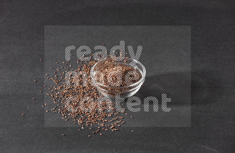 A glass bowl full of flaxseeds surrounded by the seeds on a black flooring