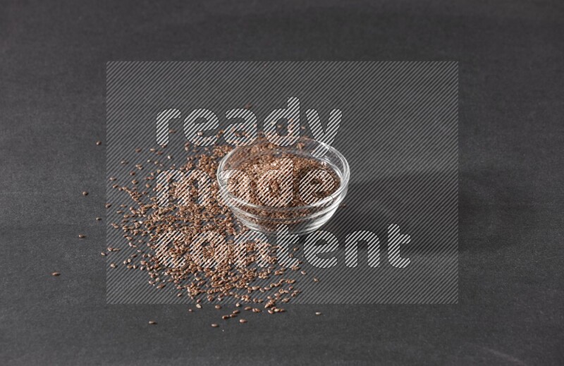 A glass bowl full of flaxseeds surrounded by the seeds on a black flooring