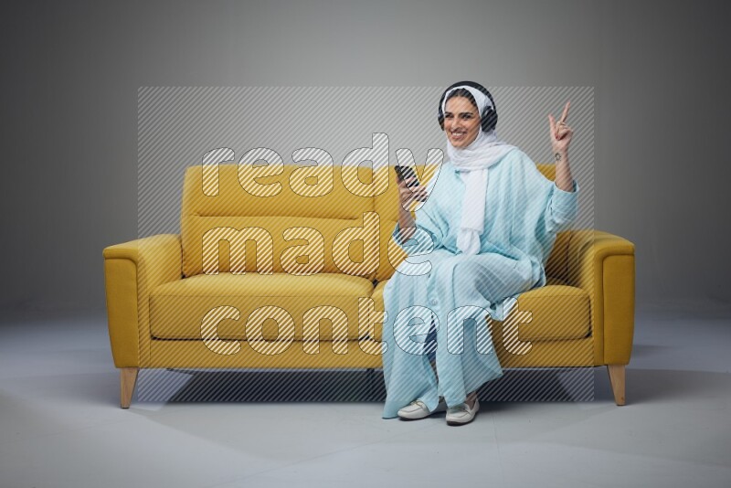 A Saudi woman wearing a light blue Abaya and a white head scarf sitting on a yellow sofa and holding an iPad while wearing headphone eye level on a grey background