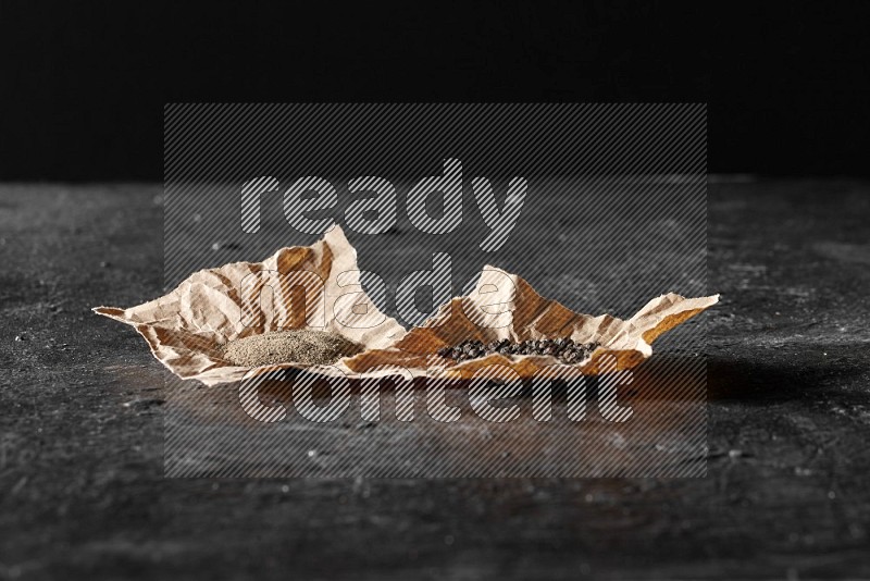 Black pepper and black pepper powder on 2 crumpled paper on a textured black flooring