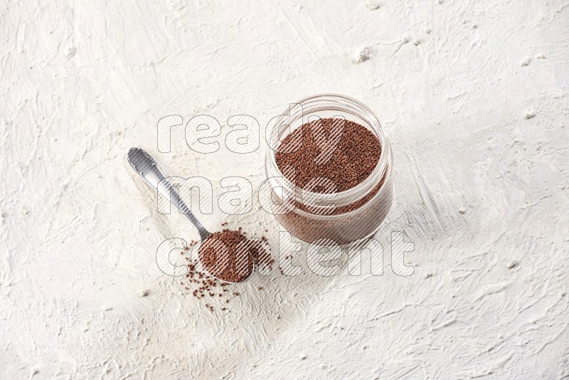 A glass jar and a metal spoon full of garden cress on a textured white flooring in different angles