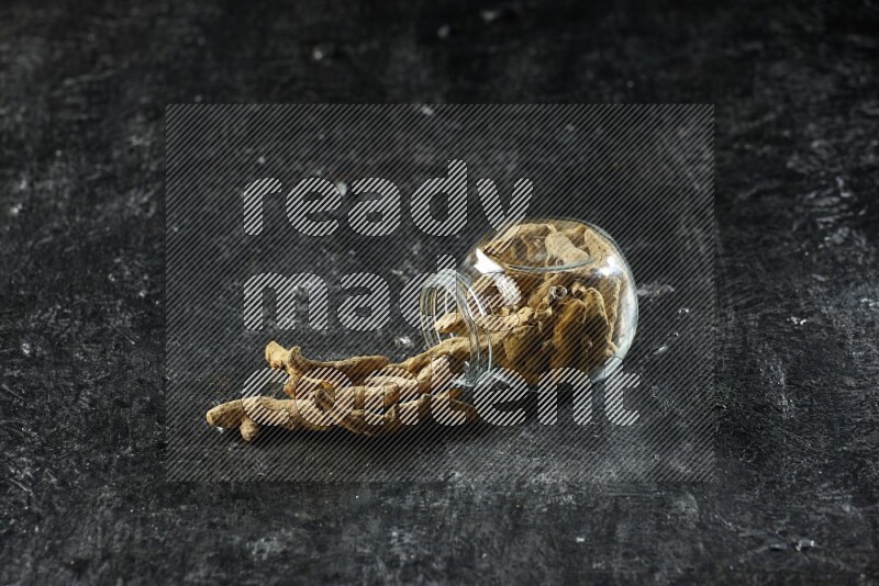 وعاء زجاجي للتوابل مقلوب وممتلئ بأصابع الكركم المجففة وبعض أصابع الكركم خارج الوعاء على خلفية سوداء