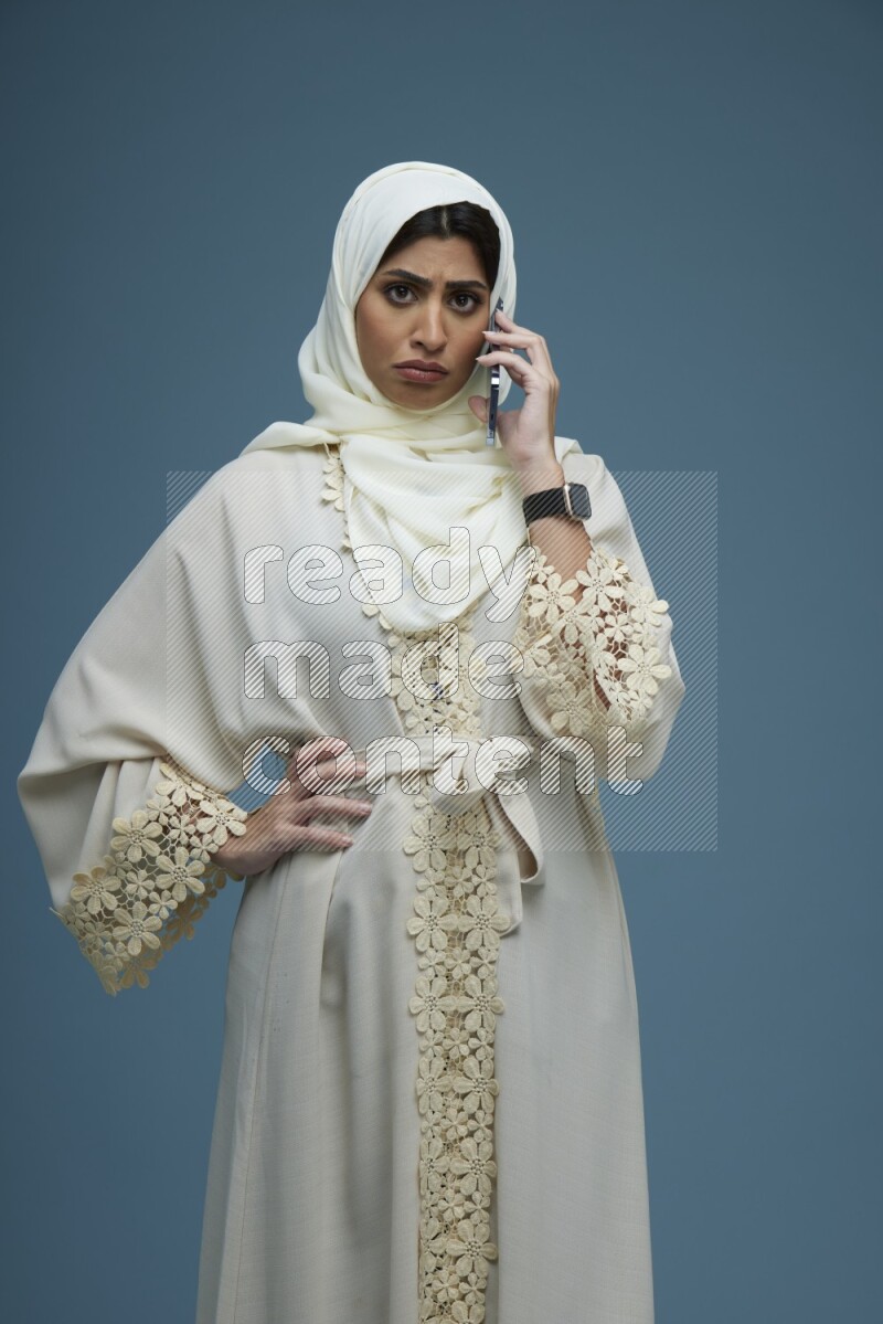 A Saudi woman having a call in a blue background wearing an off-white Abaya Hijab