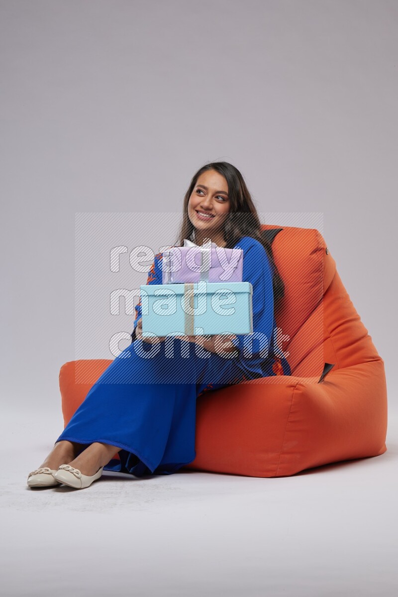 A woman sitting on an orange beanbag wearing Jalabeya holding a gift box