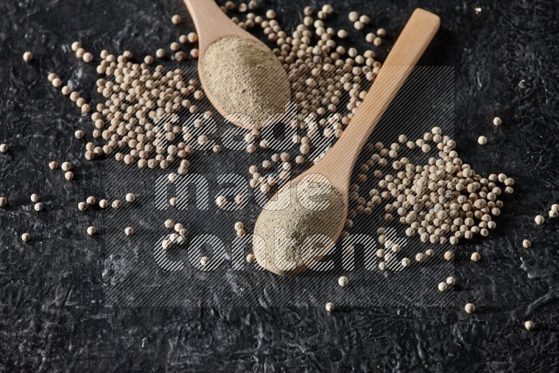 2 wooden spoons full of white pepper powder with white pepper beads on textured black flooring