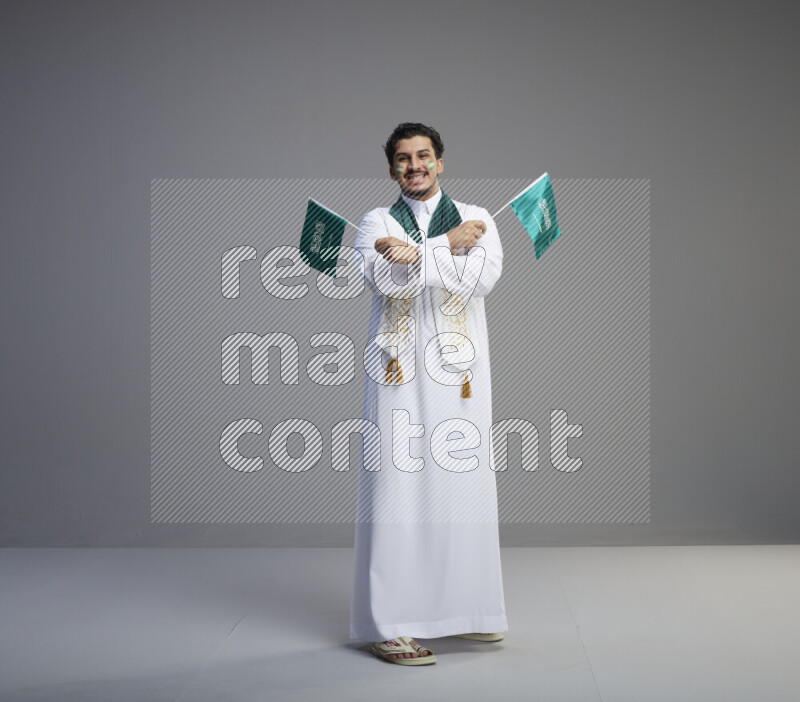 A Saudi man standing wearing thob and saudi flag scarf with face painting holding small Saudi flag on gray background