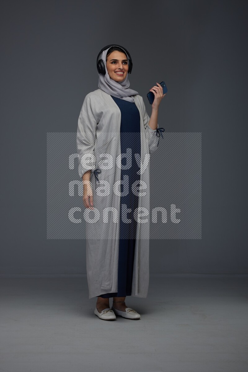 A Saudi woman wearing a light gray Abaya and head scarf standing and listening to music on a grey background