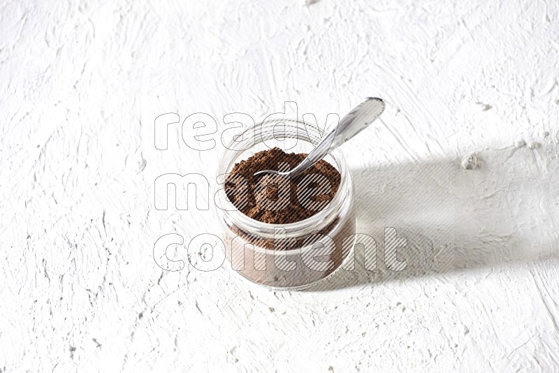 A glass jar full of cloves powder with a metal spoon on a textured white flooring