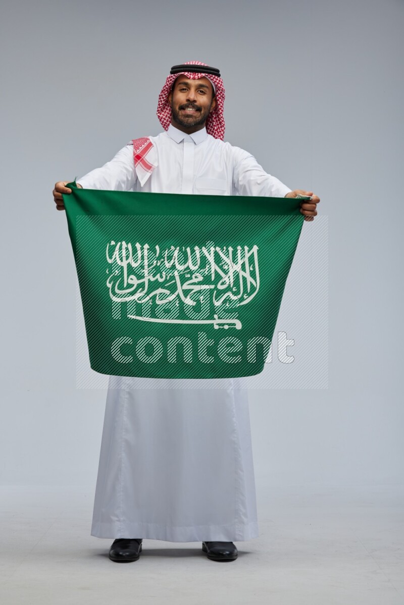 Saudi man Wearing Thob and shomag standing holding Saudi flag on Gray background