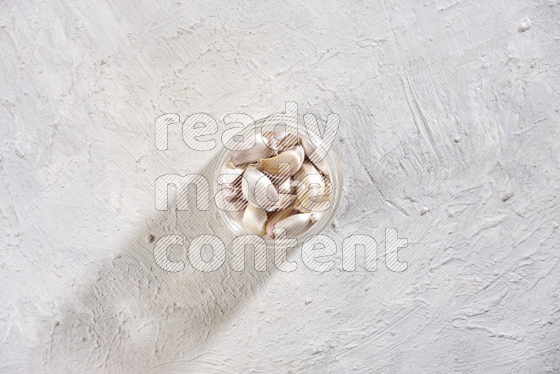 A glass jar full of garlic cloves on a textured white flooring in different angles
