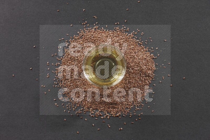 A glass bowl full of flaxseeds oil surrounded by the seeds on a black flooring