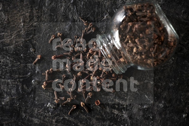 A flipped glass spice jar full of cloves and cloves came out of it on textured black flooring