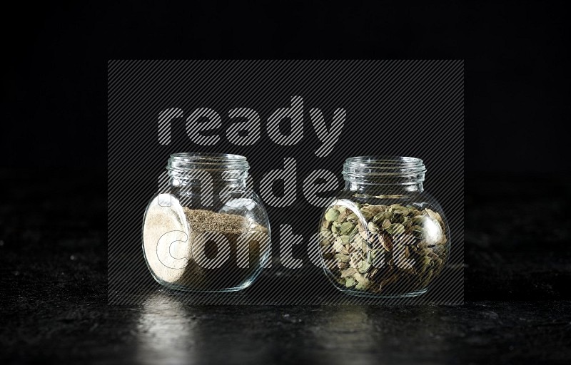 2 glass spice jars full of cardamom powder and cardamom seeds on textured black flooring