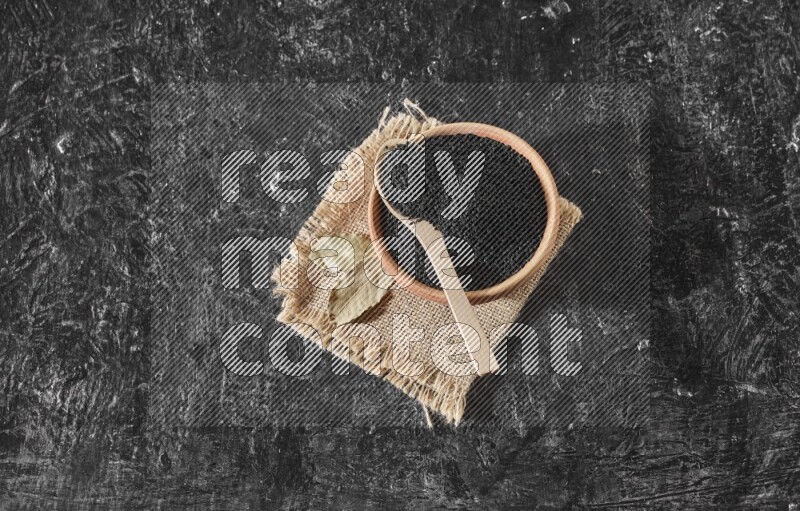 A wooden bowl full of black seeds with wooden spoon full of the seeds on it on a burlap fabric on a textured black flooring