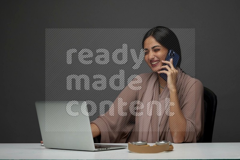 A Saudi woman Sitting on her desk Calling  on a Gray Background wearing Brown Abaya