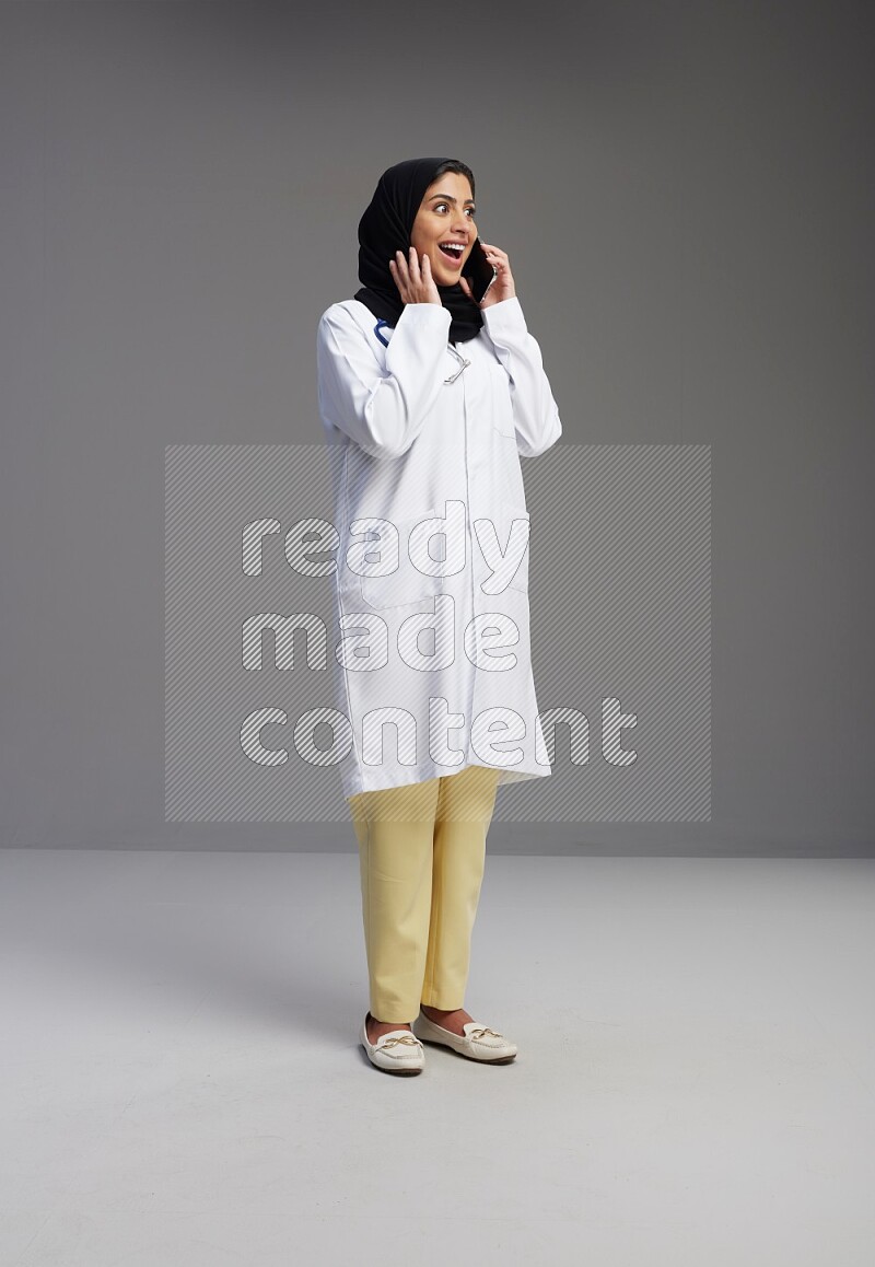 Saudi woman wearing lab coat with stethoscope standing talking on phone on Gray background