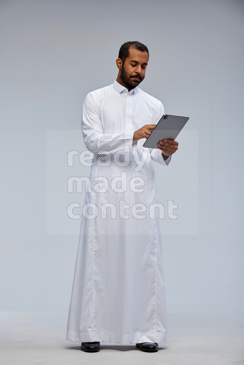 Saudi man wearing Thob standing working on tablet on Gray background