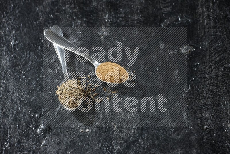 ملعقتان معدنيتان مملوءتان بحبوب الكمون وبودرة الكمون على أرضية سوداء