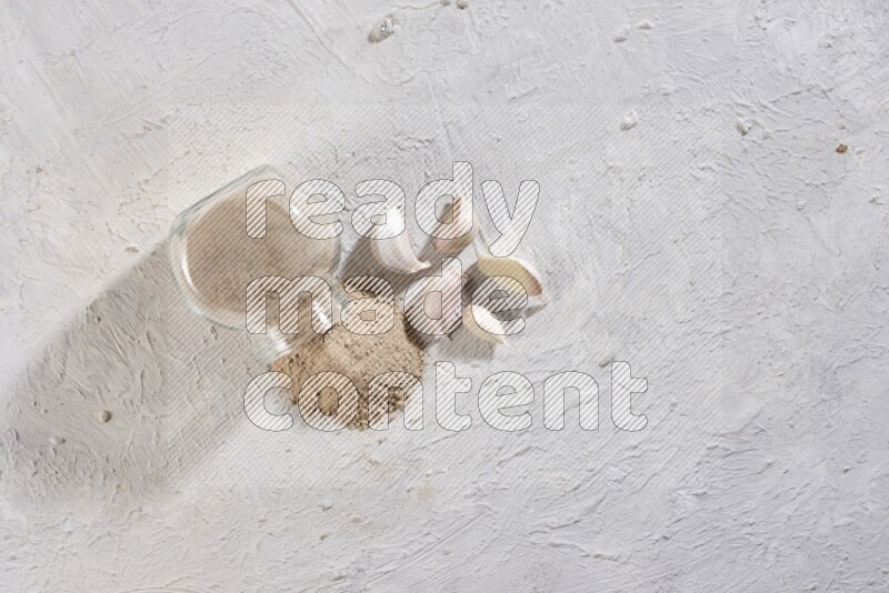 وعاء توابل زجاجي ممتلئ ببودرة الثوم مقلوب والبودرة متناثرة منه علي خلفية بيضاء