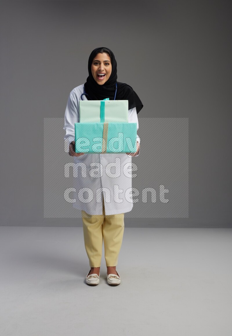 Saudi woman wearing lab coat with stethoscope standing holding gift box on Gray background