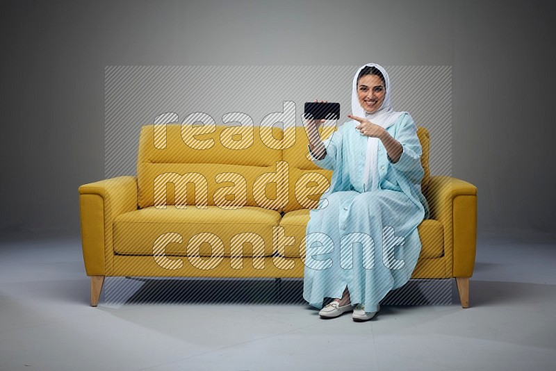 A Saudi woman wearing a light blue Abaya and white head scarf sitting on a yellow sofa and showing her phone's screen vertically and horizontally eye level on a grey background