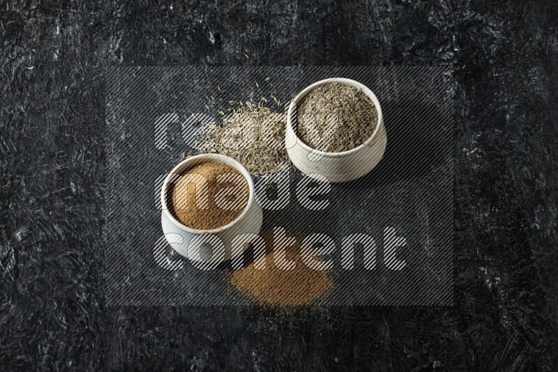 2 beige bowls, one full of cumin powder and the other full of seeds on a textured black flooring