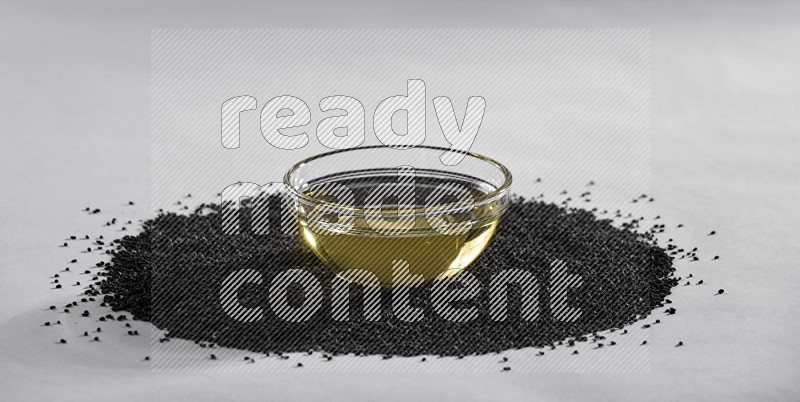 A glass bowl full of black seeds oil surrounded by the seeds on a white flooring