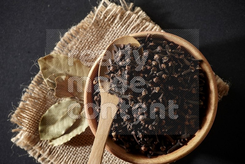 A wooden bowl, a wooden spoon full of cloves, and bay leaves (laurel) on a piece of burlap on a black flooring
