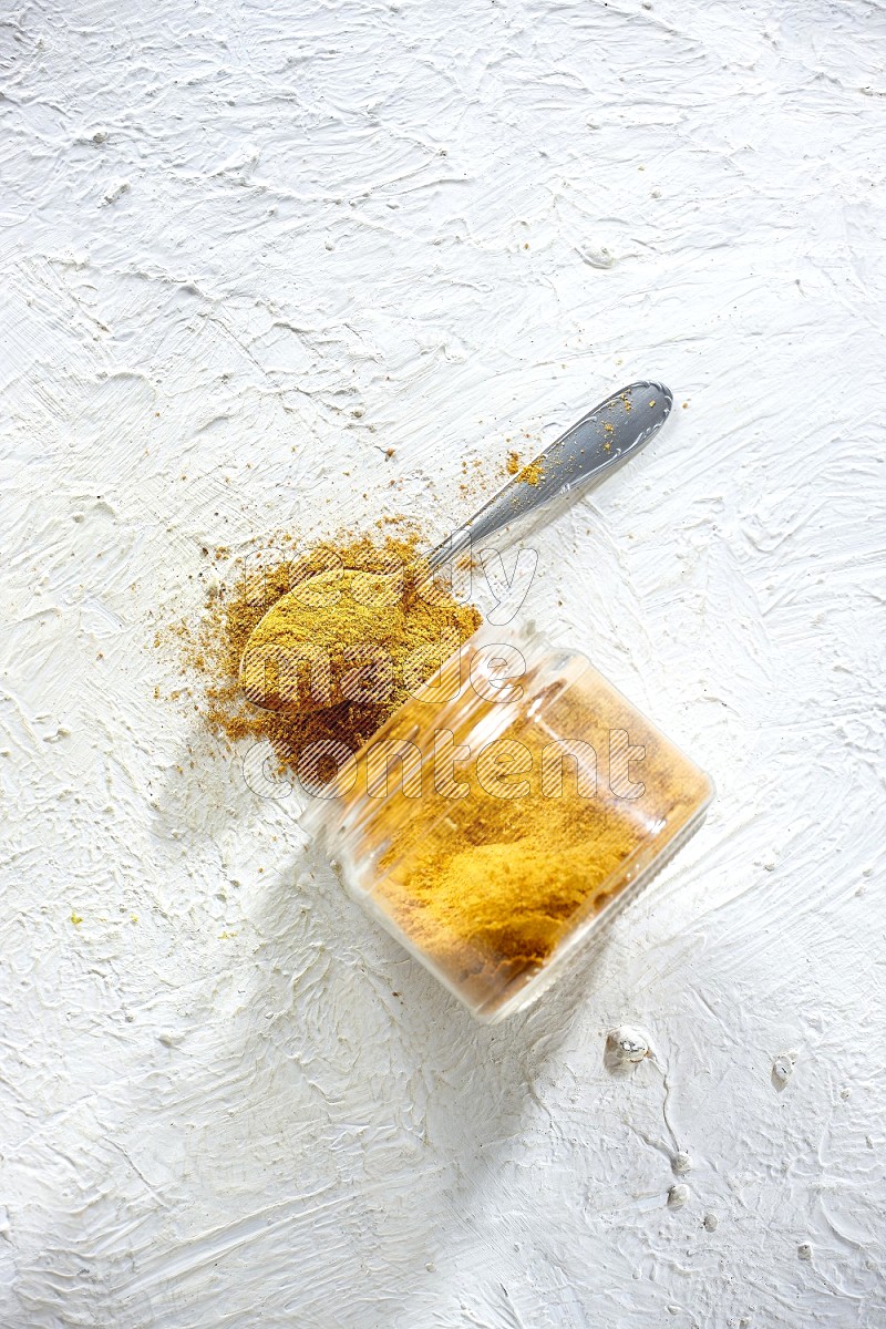 A flipped glass spice jar and a metal spoon full of turmeric powder and powder spilled out of it on textured white flooring