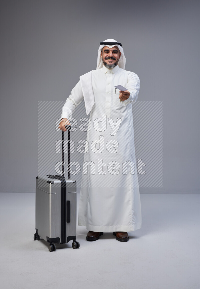Saudi man wearing Thob and white Shomag standing holding Travel bag and ATM card on Gray background