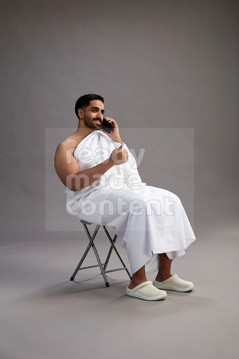 A man wearing Ehram sitting on chair talking on the phone on gray background
