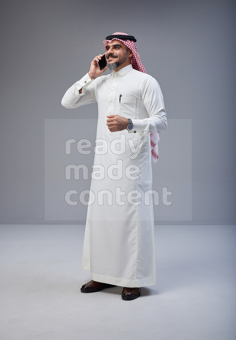 Saudi man Wearing Thob and red Shomag standing talking on phone on Gray background