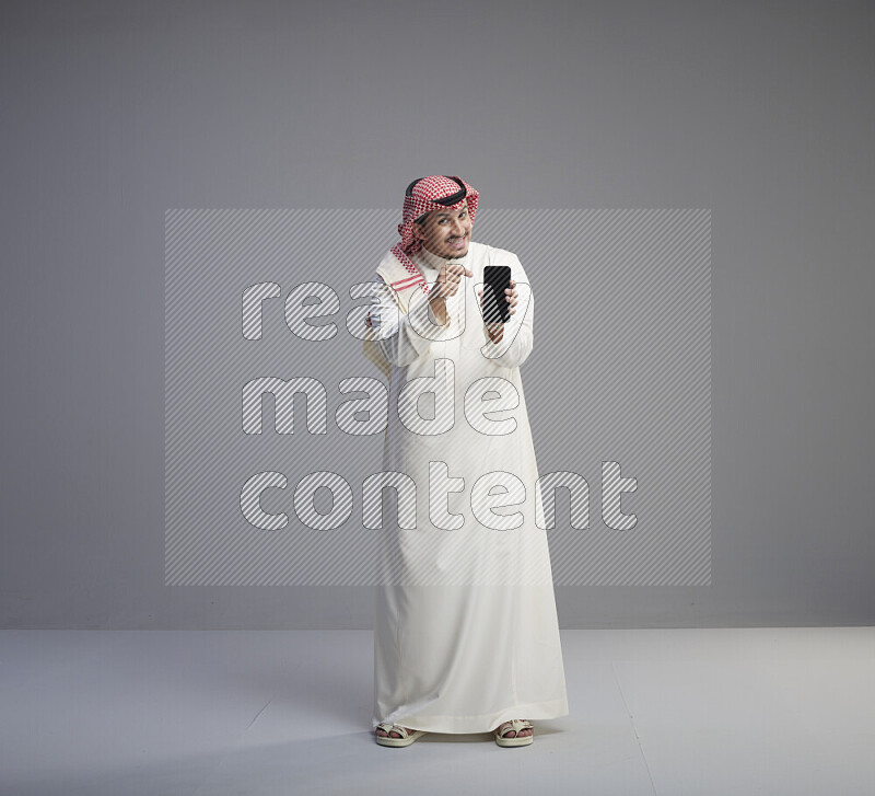 رجل سعودي يرتدي ثوب ابيض وشماغ احمر يشير الى هاتفه باتجاه الكاميرا