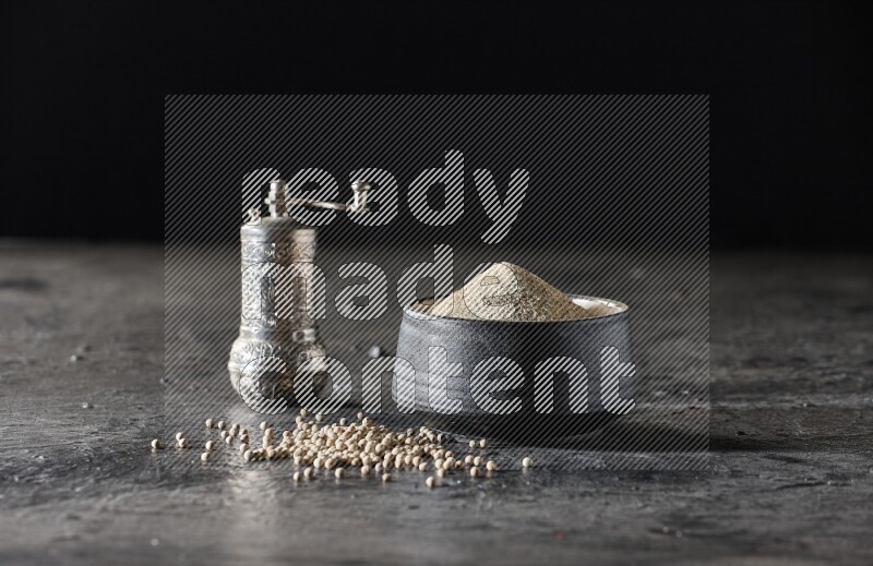 وعاء فخار أسود مملوء ببودرة الفلفل الأبيض مع حبات الفلفل ومطحنة الفلفل المعدنية على أرضية سوداء