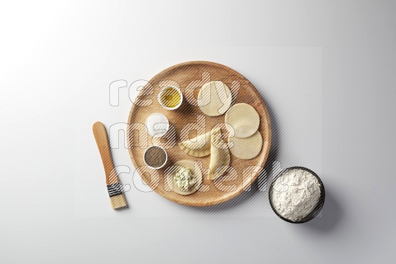 two closed sambosas and one open sambosa filled with cheese while flour, salt, black pepper and oil with oil brush aside in a wooden dish on a white background