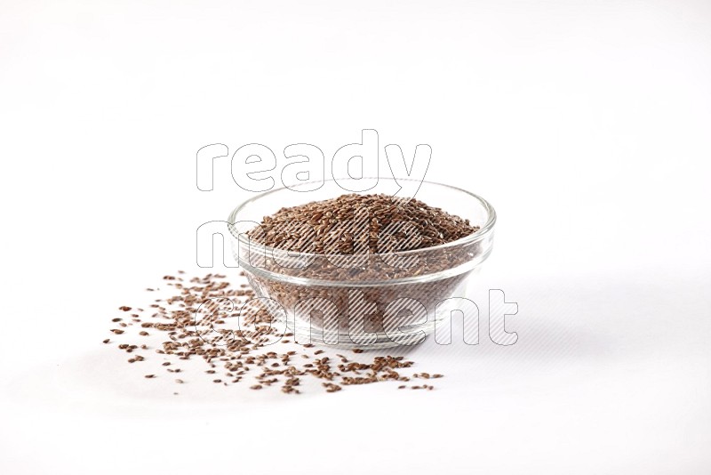 A glass bowl full of flax seeds surrounded by flax seeds on a white flooring