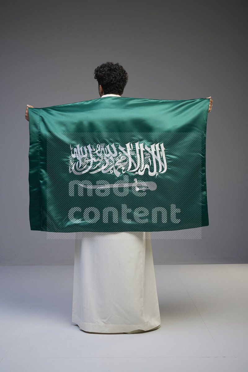 A saudi man standing wearing thob holding big saudi flag on gray background