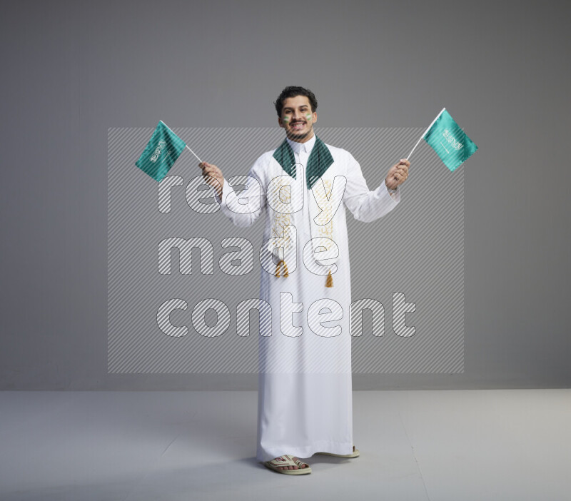 A Saudi man standing wearing thob and saudi flag scarf with face painting holding small Saudi flag on gray background