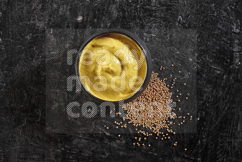 A black pottery bowl full of mustard paste with mustard seeds underneath on textured black flooring in different angles