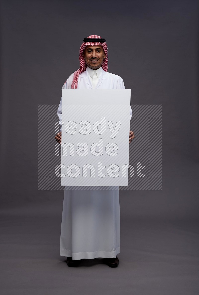 Saudi man wearing thob with lab coat and shomag with pocket employee badge standing holding board on gray background