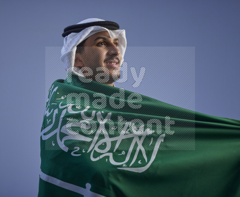 A close-up shot of Saudi man wearing thob and white shomag wrapping big Saudi flag on gray background