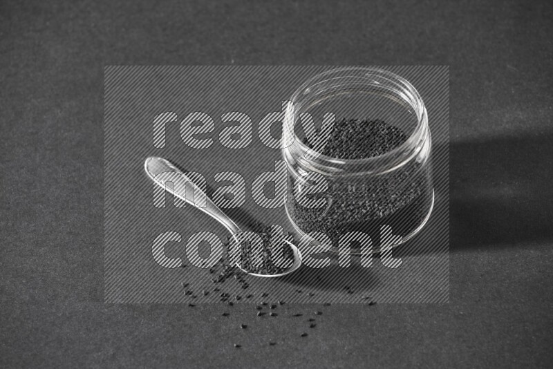 A glass jar full of black seeds with a metal spoon full of the seeds on a black flooring