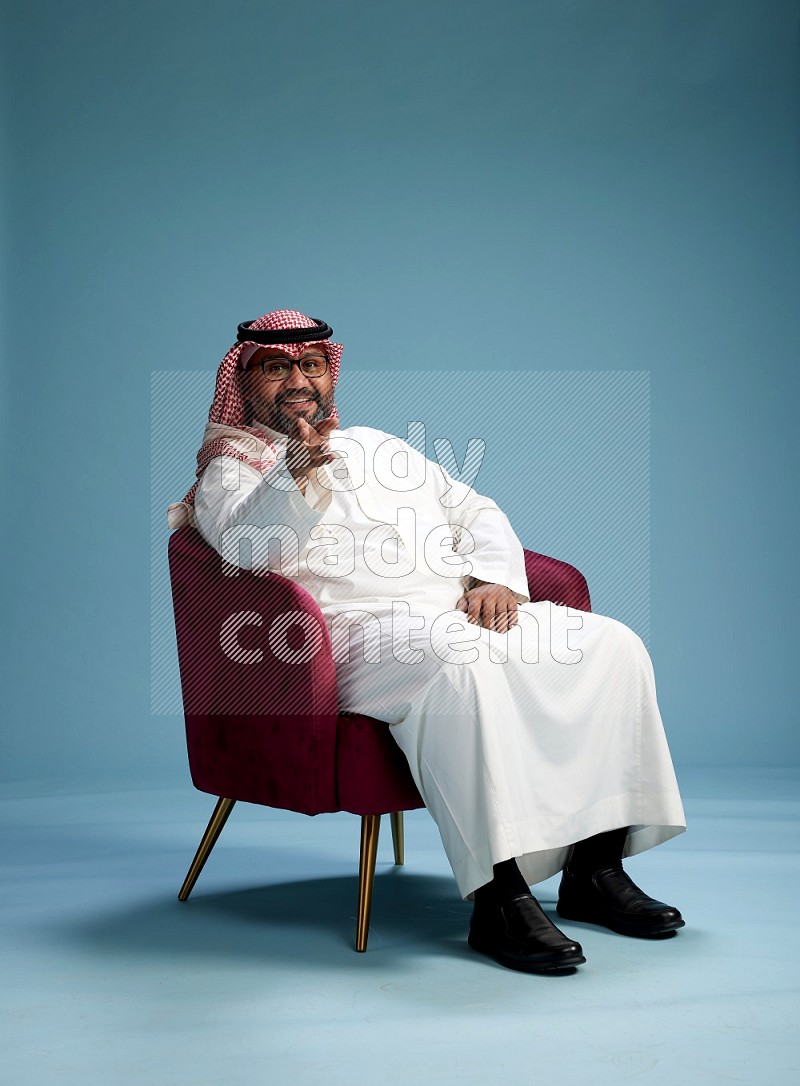 Saudi Man with shimag sitting on chair Interacting with the camera on blue background