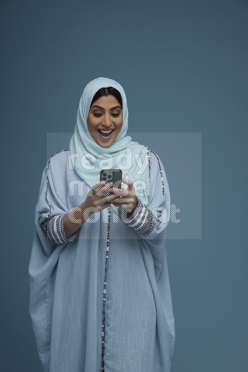 A Saudi woman Texting in a blue background wearing a blue Abaya with hijab