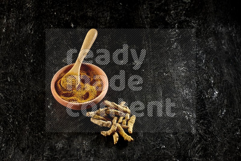 A wooden bowl and a wooden spoon full of turmeric powder with dried turmeric fingers on textured black flooring