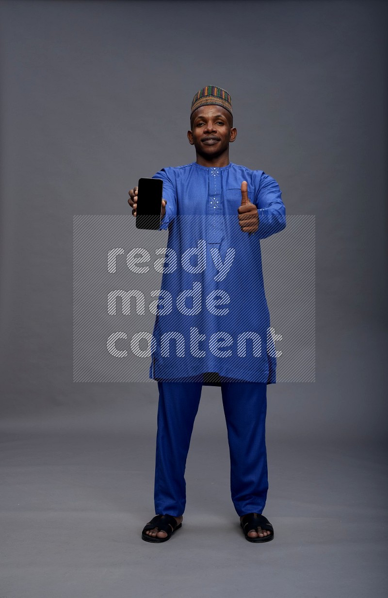 Man wearing Nigerian outfit standing showing phone to camera on gray background