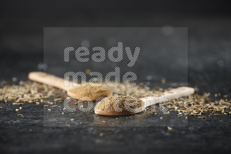 ملعقتان خشبيتان ممتلئتان ببودرة الكمون مع حبوب الكمون المتناثرة على خلفية سوداء