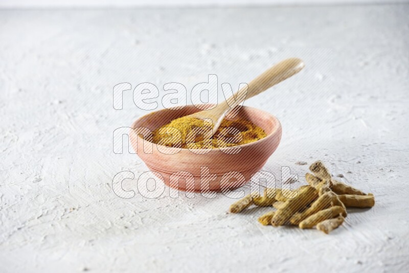 A wooden bowl and wooden spoon full of turmeric powder with dried turmeric fingers beside it on textured white flooring