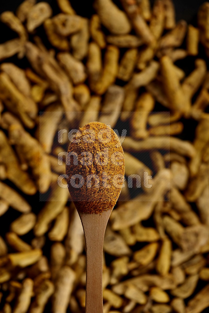 A wooden spoon full of turmeric powder above dried turmeric fingers on black flooring