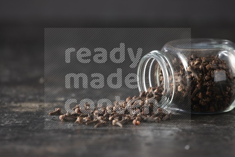 A flipped glass spice jar full of cloves and cloves came out of it on textured black flooring
